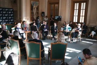 Oamenii stau așezați în scaune pe o platformă în fața unui panou negru, participând la o discuție într-o încăpere cu pereți înalți și ferestre mari.