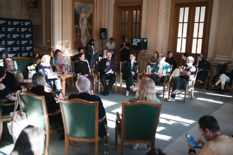 Oamenii stau așezați în scaune pe o platformă în fața unui panou negru, participând la o discuție într-o încăpere cu pereți înalți și ferestre mari.