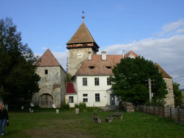 O clădire veche cu turn înalt și acoperiș din țiglă roșie este situată pe un teren cu iarbă verde sub un cer albastru.