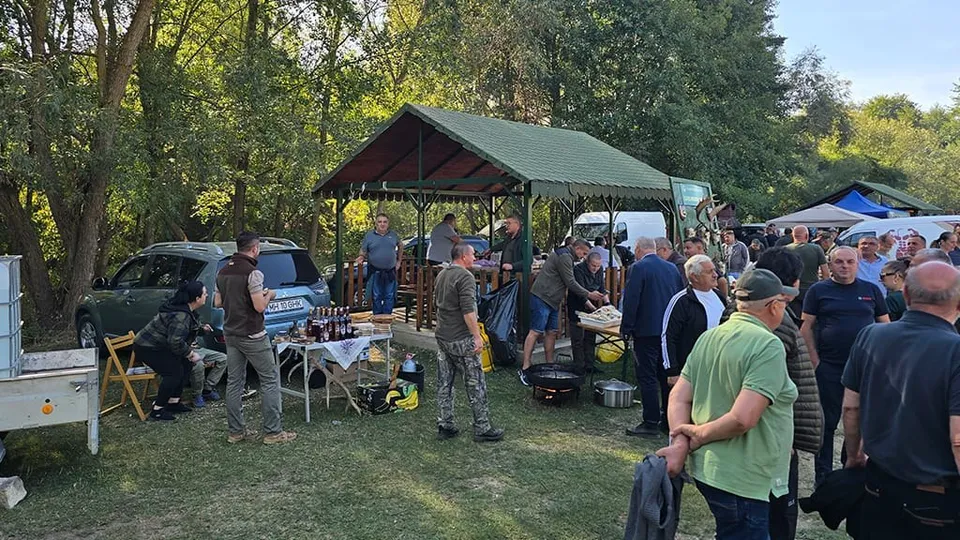 Un grup de oameni stă pe un teren verde lângă un pavilion cu acoperiș verde, mese cu obiecte și mașini parcate în fundal.
