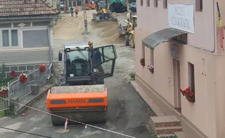 Un aparat de compactare portabil de culoare portocalie și neagră este poziționat pe un drum de pietriș între două clădiri.