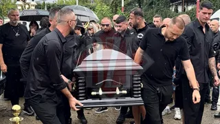 Un grup de bărbați îmbrăcați în negru poartă un sicriu întunecat printr-o mulțime de oameni în aer liber.