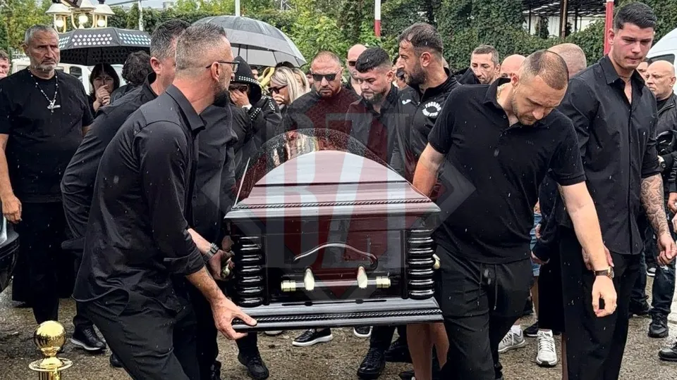 Un grup de bărbați îmbrăcați în negru poartă un sicriu întunecat printr-o mulțime de oameni în aer liber.