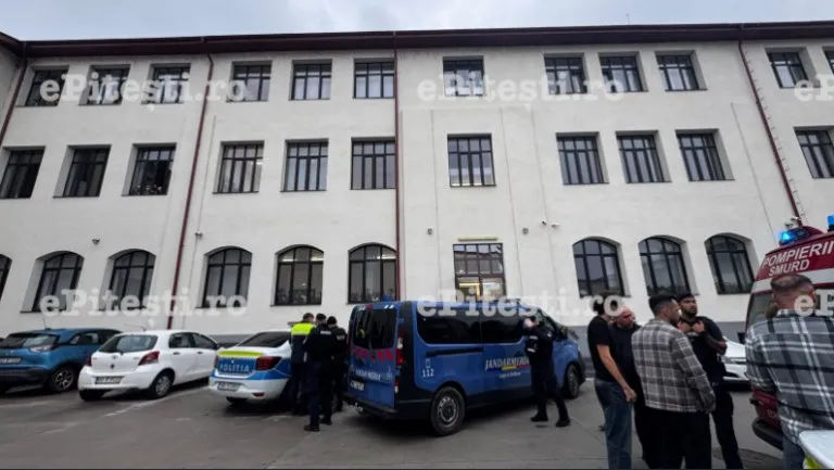 Mai mulți oameni stau lângă mașini de poliție și ambulanță parcate în fața unei clădiri albe cu ferestre multiple.