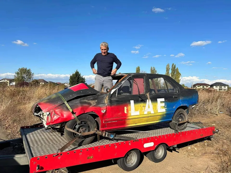 Un bărbat stă în picioare lângă o mașină roșu-albastră cu partea frontală zdrobită, aflată pe o platformă de transport roșie sub un cer senin.