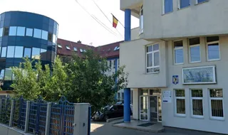 O clădire albă cu ferestre și un imobil curbat cu fațadă de sticlă albastră sunt vizibile în spatele unui gard metalic albastru.