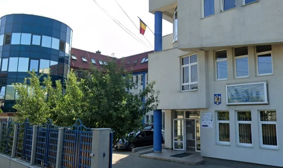 O clădire albă cu ferestre și un steag este situată lângă un imobil curbat cu fațadă de sticlă și un gard albastru.