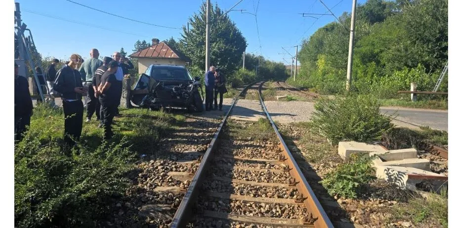 Un autoturism negru este parcat lângă șinele de feroviar, în timp ce un grup de persoane stă în apropiere pe un teren cu vegetație verde.