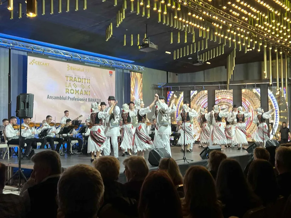 Un grup de dansatori în costume tradiționale albe cu detalii colorate execută un dans pe o scenă iluminată în fața unui public.