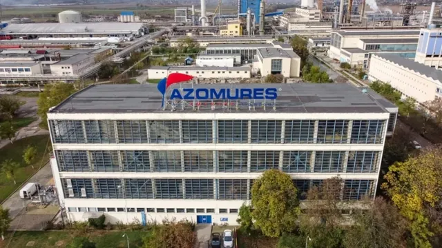O clădire de birouri cu ferestre mari și un panou publicitar albastru pe acoperiș este situată într-o zonă industrială cu diverse facilități.