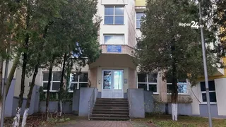 O clădire cu ferestre mari și un set de scări de acces gri este încadrată de copaci înalți pe ambele părți.