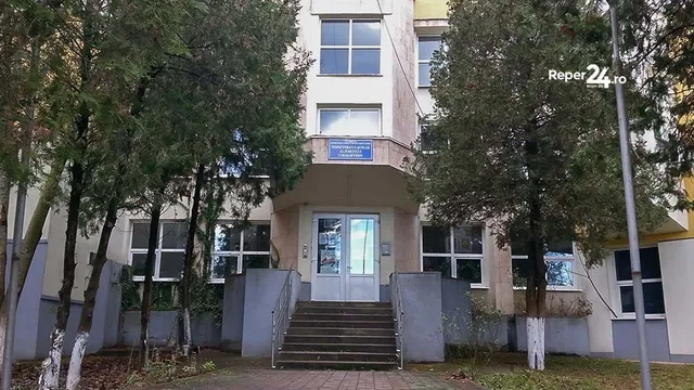 O clădire cu ferestre mari și un set de scări de acces gri este încadrată de copaci înalți pe ambele părți.