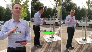 Un bărbat într-o cămașă deschisă la guler stă lângă un copac tânăr protejat de o structură metalică pe un trotuar.