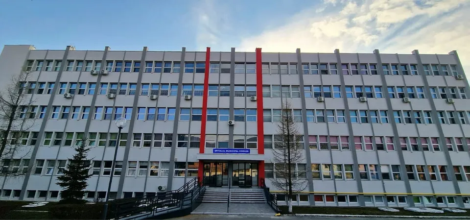 O clădire gri cu ferestre multiple și detalii verticale roșii prezintă o intrare cu scări și o rampă de acces.