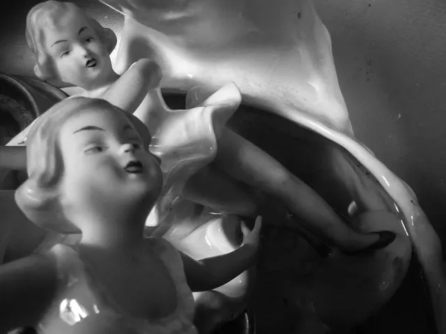 Două figurine de porțelan cu trăsături feminine sunt așezate într-o compoziție cu materiale albe și lucioase pe un fundal închis.