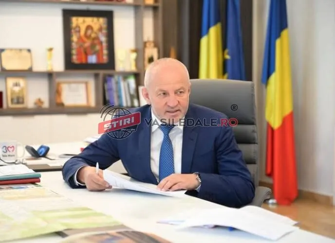 Un bărbat cu părul scurt și costum albastru stă la un birou cu hărți și documente, având steaguri și un tablou în fundal.
