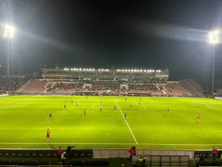 Jucători de fotbal sunt împrăściați pe un teren verde sub lumini puternice de stadion, în fața unor tribune cu spectatori.