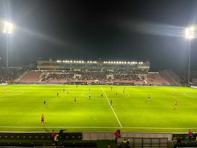 Jucători de fotbal în echipamente colorate sunt dispersați pe un teren verde de sport sub reflectoarele unui stadion mare pe timp de noapte.