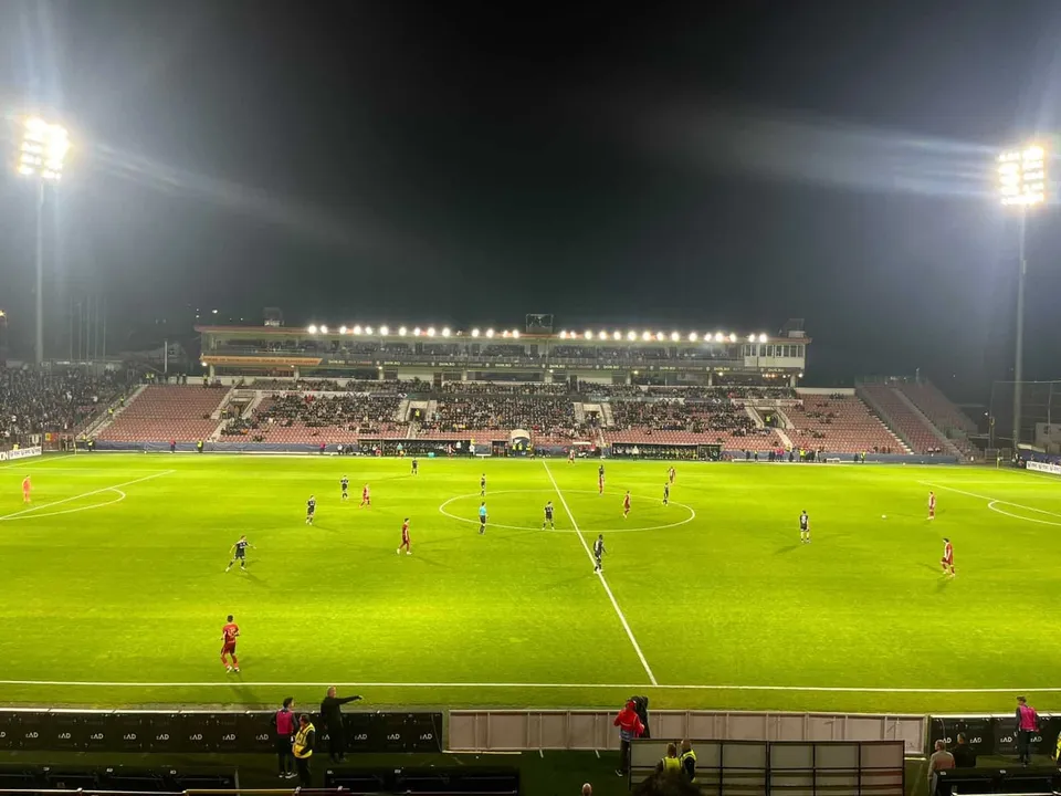 Jucători de fotbal în echipamente colorate sunt dispersați pe un teren verde de sport sub reflectoarele unui stadion mare pe timp de noapte.