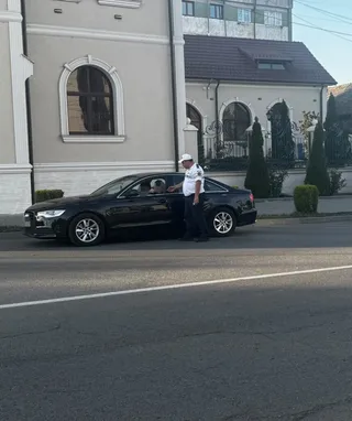 Un agent de poliție în uniformă albă și pantaloni închiși stă lângă o mașină neagră pe marginea unui drum asfaltat.