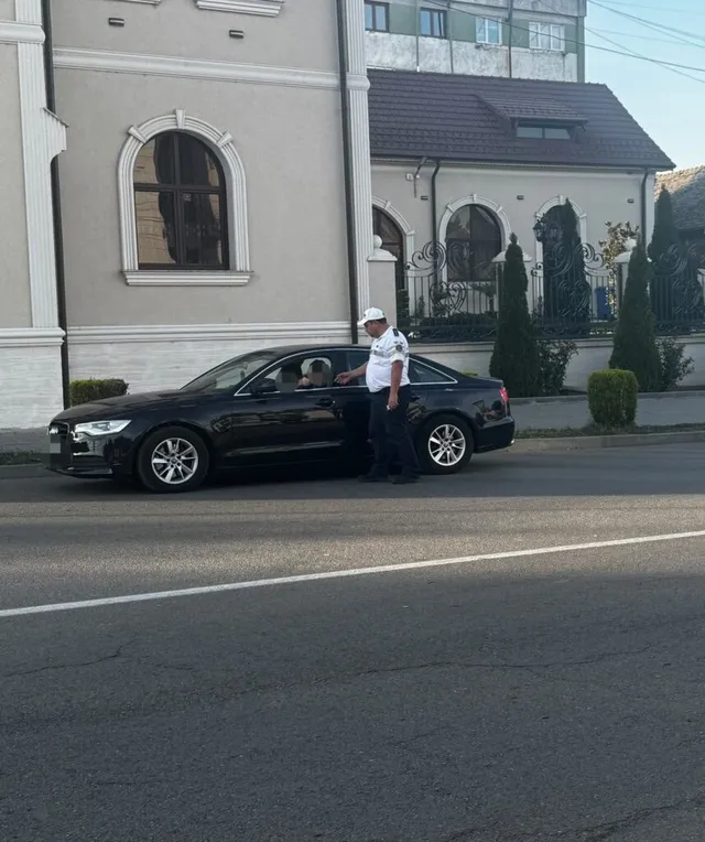 Un agent de poliție în uniformă albă și pantaloni închiși stă lângă o mașină neagră pe marginea unui drum asfaltat.