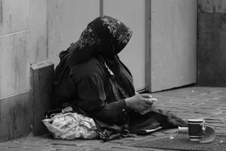 O persoană cu un basma înflorită stă așezată pe trotuar lângă un perete, având o geantă și o cană lângă ea.