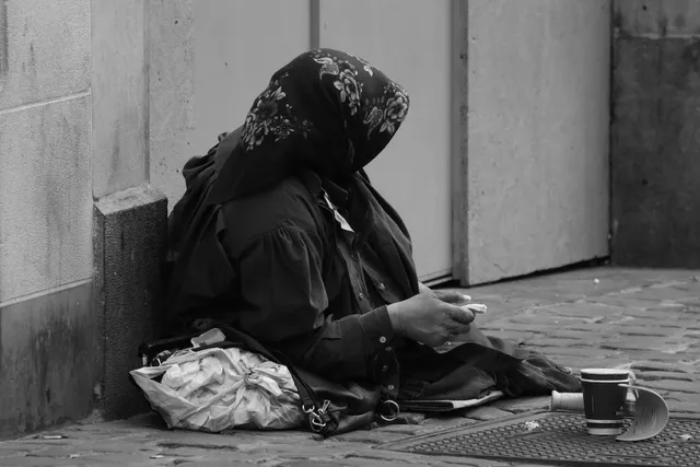 O persoană cu un basma înflorită stă așezată pe trotuar lângă un perete, având o geantă și o cană lângă ea.