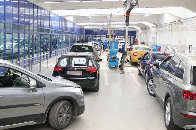 Mai multe autoturisme sunt parcate într-un spațiu interior cu pereți albi și panouri de sticlă albastră.