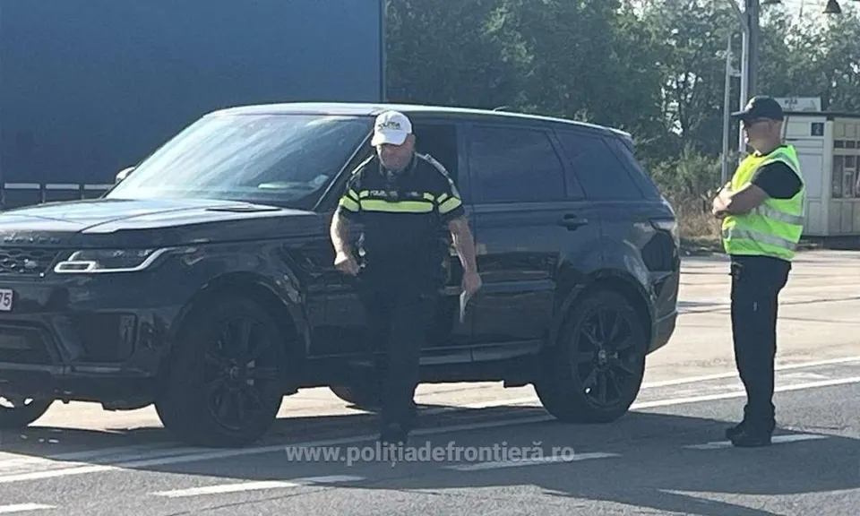 Un agent de poliție în uniformă și vestă reflectorizantă stă lângă o mașină neagră, în timp ce un alt agent se află în partea dreaptă.