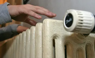 O mână întinsă deasupra unui calorifer metalic de culoare deschisă dotat cu un termostat alb.