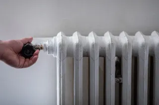 O mână întinde degetele spre un robinet negru de pe un radiator alb de metal.