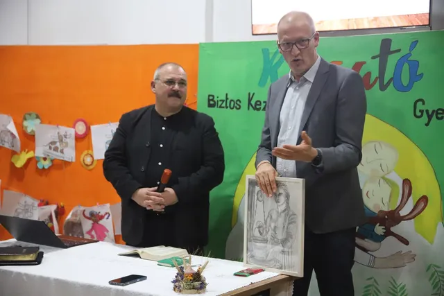 Un bărbat cu ochelari și sacou gri vorbește lângă un tablou, în timp ce un alt bărbat cu haine negre stă în spatele unei mese.