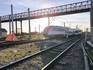 Un tren modern de culoare albă circulă pe o cale ferată sub o pod de metal și cabluri electrice suspendate.