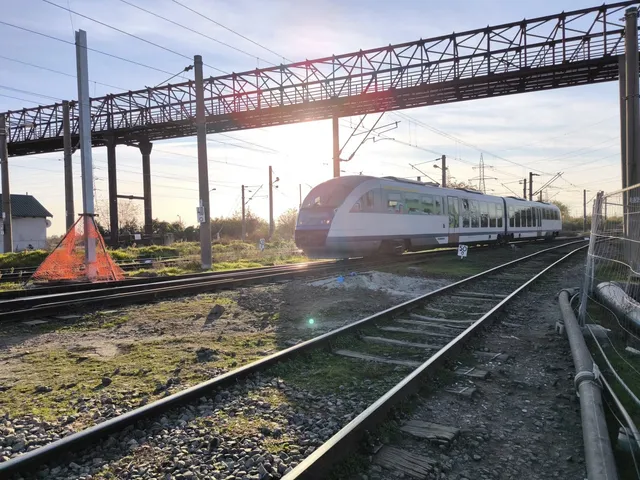 Un tren modern de culoare albă circulă pe o cale ferată sub o pod de metal și cabluri electrice suspendate.