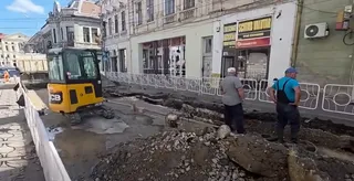 Un utilaj de construcții galben și negru stă pe un drum parțial săpat lângă clădiri urbane, în prezența a doi bărbați care privesc spre șantier.