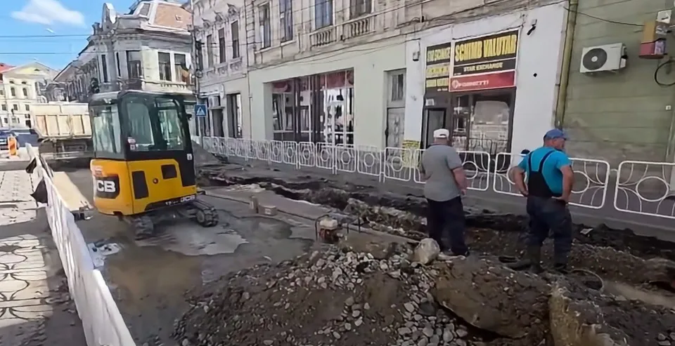 Un utilaj de construcții galben și negru stă pe un drum parțial săpat lângă clădiri urbane, în prezența a doi bărbați care privesc spre șantier.