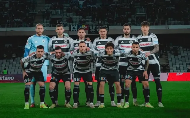 O echipă de fotbaliști în echipamente alb-negre stă în poziție de grup pe un teren de fotbal, cu câțiva jucători în picioare și alții așezați.