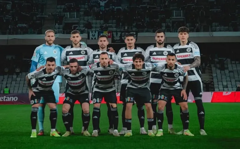 O echipă de fotbaliști în echipamente alb-negre stă în poziție de grup pe un teren de fotbal, cu câțiva jucători în picioare și alții așezați.