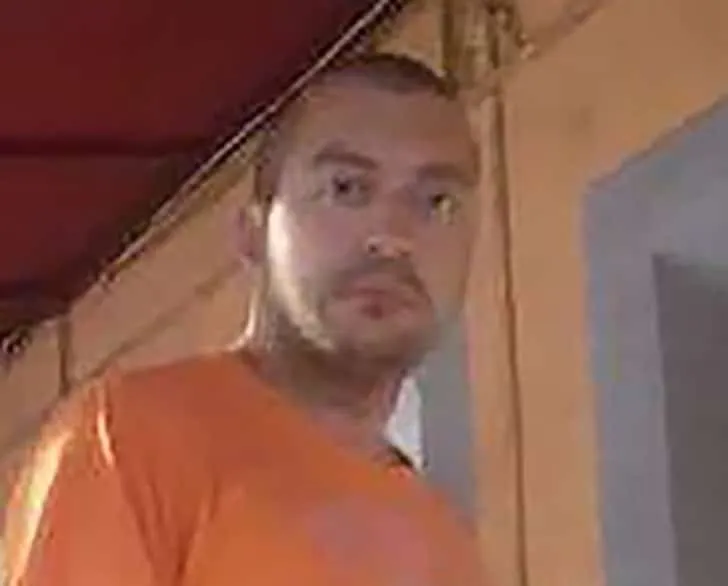 Un bărbat cu barbă scurtă și tricou portocaliu privește spre cameră într-un cadru cu lumină caldă.