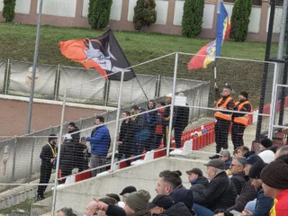 Un grup de persoane stă pe tribunele unui stadion lângă un gard metalic, sub două steaguri colorate agitate în vânt.