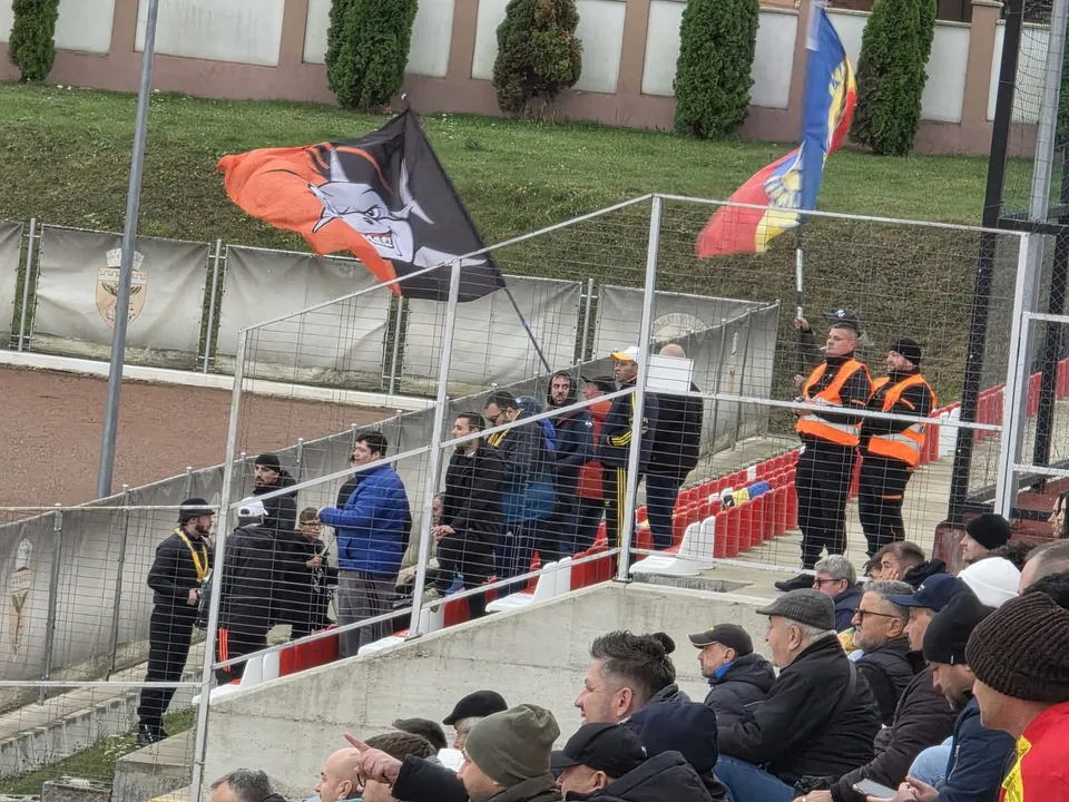 Un grup de persoane stă pe tribunele unui stadion lângă un gard metalic, sub două steaguri colorate agitate în vânt.