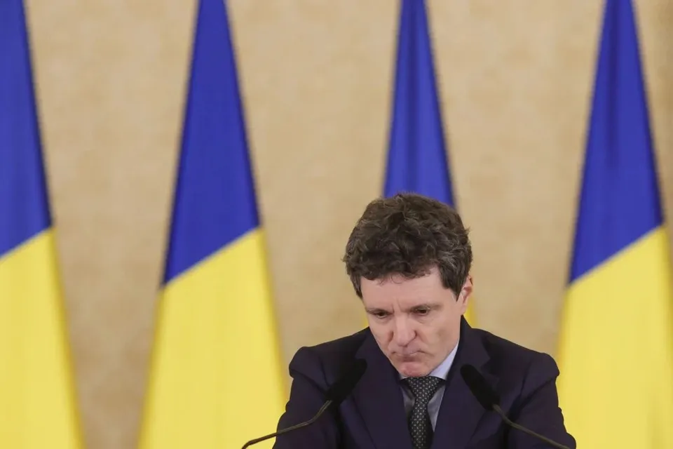 Un bărbat cu părul încrețit, purtând o jachetă închisă la culoare și cravată, stă în fața unor steaguri albastre și galbene.