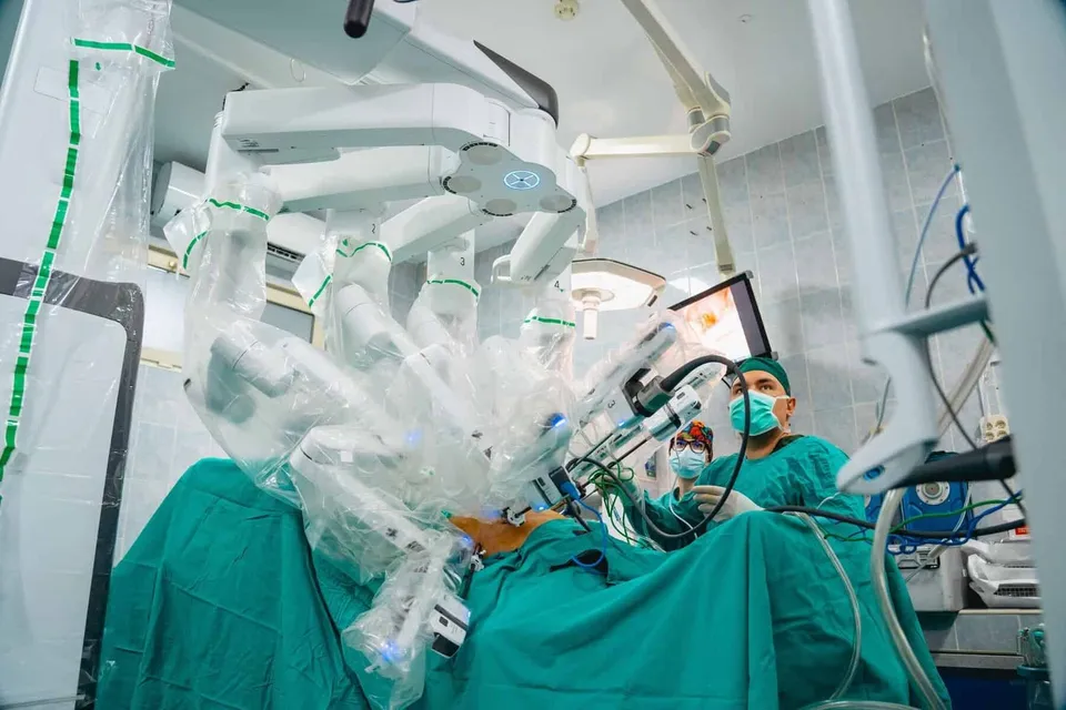 Un medic cu mască și căciulă verde operează folosind un aparat robotic acoperit cu folie transparentă într-o sală de operație.