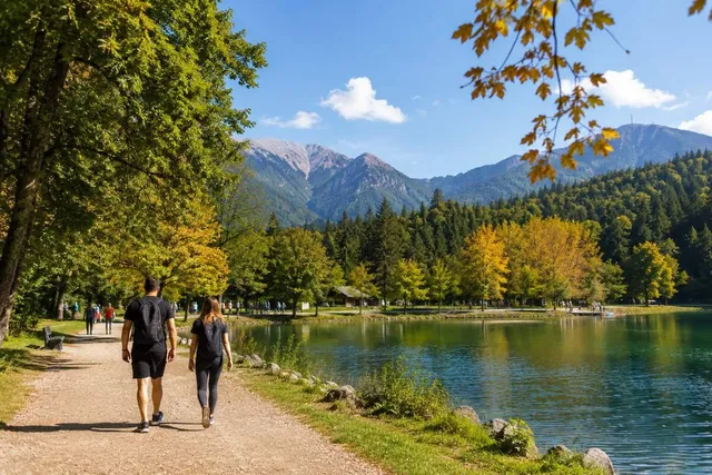 Oamenii merg pe un drum de pământ lângă un lac, înconjurați de copaci cu frunze verzi și galbene și de munți în fundal.