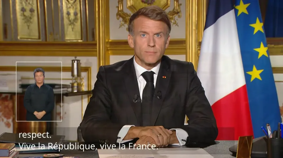 Un bărbat într-un costum negru și cravată neagră stă la o masă în fața unui steag francez și al unuia european.