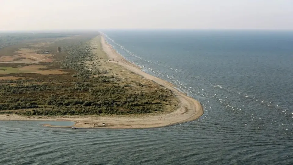 O vedere aeriană prezintă o bucată de pământ îngustă, acoperită cu vegetație, situată între o masă de apă și o coastă nisipoasă.