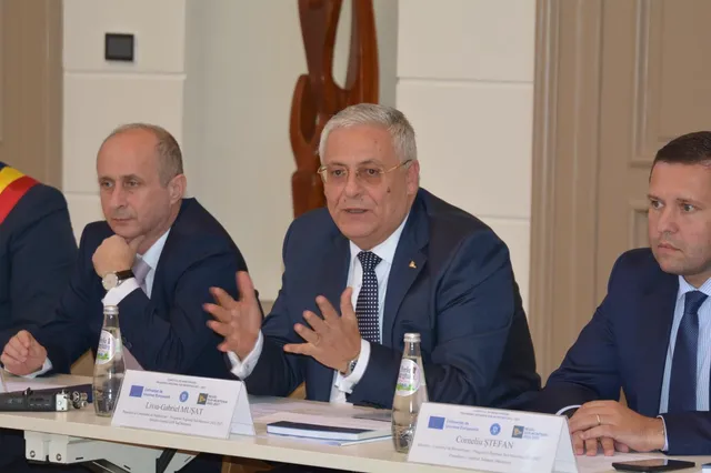 Trei bărbați în costume purtând cămăși și cravate stau la o masă albă cu sticle de apă și documente în fața unui perete deschis.