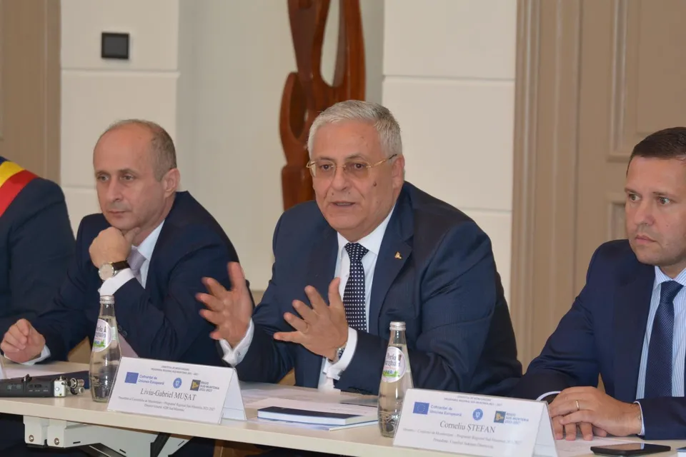 Trei bărbați în costume purtând cămăși și cravate stau la o masă albă cu sticle de apă și documente în fața unui perete deschis.