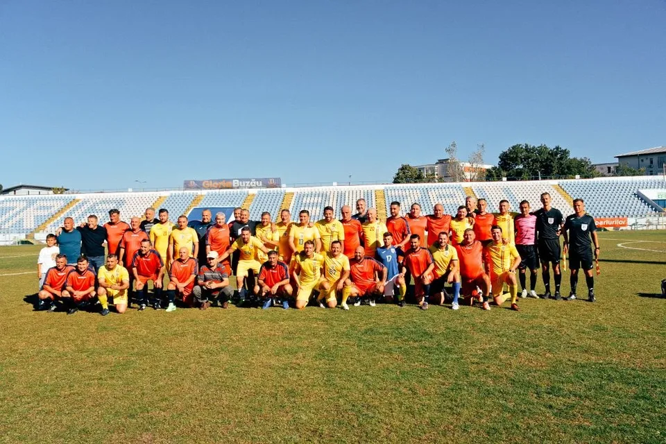 Un grup de bărbați în echipamente sportive colorate stau poziționați pe un teren de fotbal verde sub un cer senin.