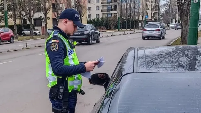 Un polițist în uniformă și vestă reflectorizantă ține documente lângă o mașină neagră pe un drum urban cu clădiri și alte vehicule în fundal.